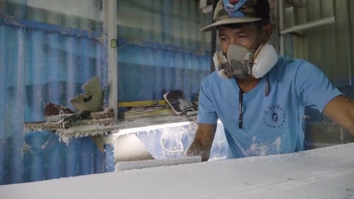 Asian man with respirator on sanding a DIY foam surfboard in workshop