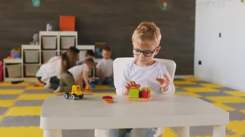 White shirt, eyewear, sitting by table and playing. Group of children are in the kindergarten.