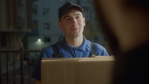 Attractive young delivery man courier in blue cap smiling into customer is delivering boxes around