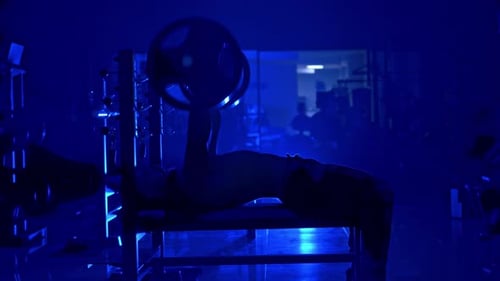 Handsome young man doing bench press workout in gym. Blue light filter. Strong abs. Heavy lift.