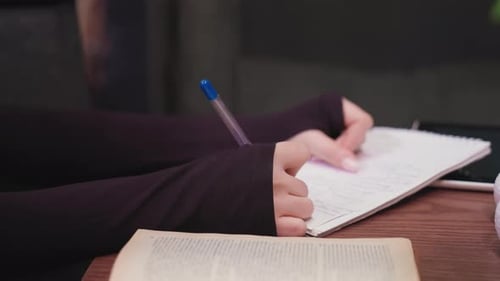 Close Up Of Woman Writing In Notebook With Book And White Decoration On Wooden Table