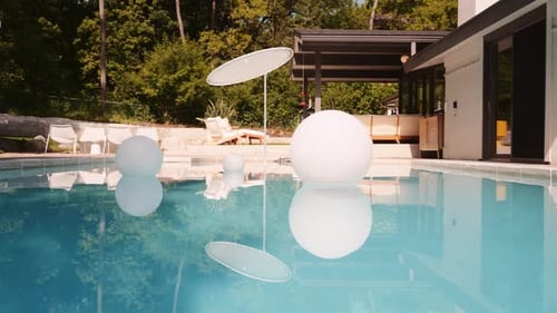 panning shot of a pool outside of a luxury home with three large floating white balls in the pool