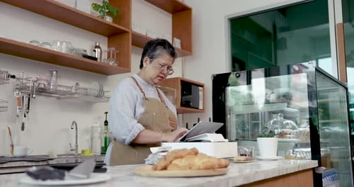 Asian senior female cafe owner looking at digital screen behind counter with serious expression
