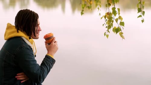 Young Handsome Man with Dreadlocks in Warm Clothes Drinking Coffee Near Lake in Golden Autumn