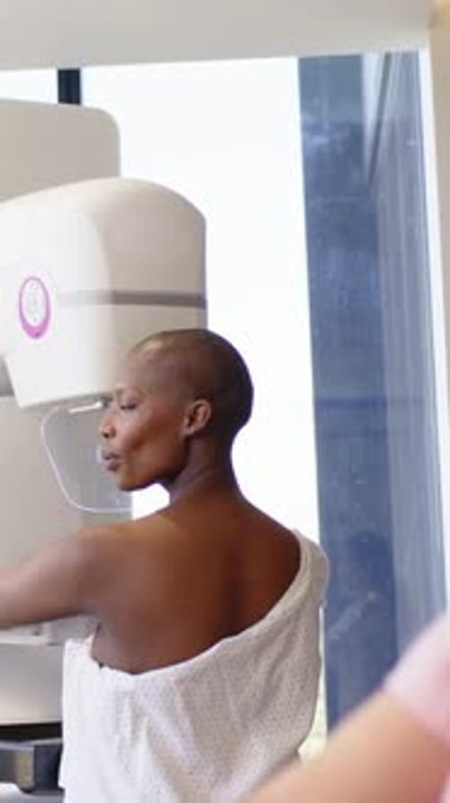 Vertical video: Tech adjusting mammogram unit, gowned woman holding handles in clinic for mammogram