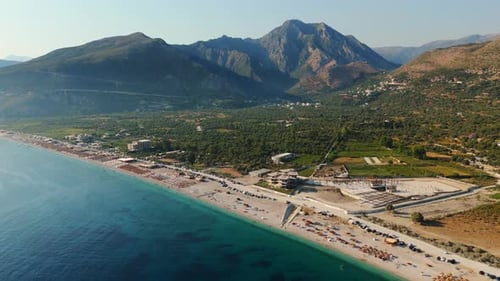 A serene beach with clear green water, scattered tourists, and mountains in the background, aerial v