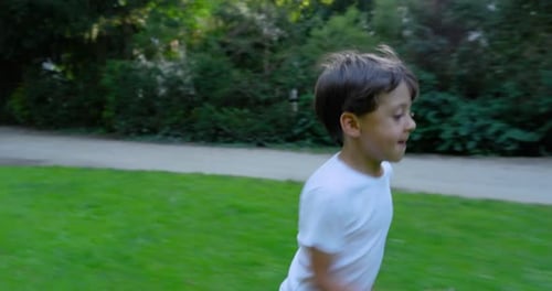Young boy running joyfully through a grassy park, wearing a white shirt, with a happy expression,