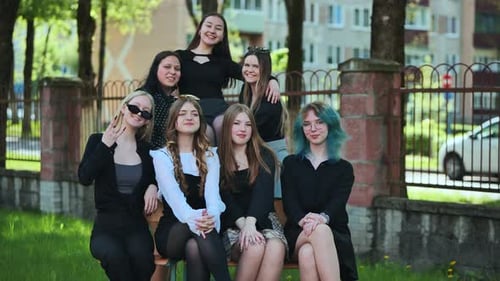 Grupo de sete moças elegantes se reúne em um banco do lado de fora da escola, tomando banho no