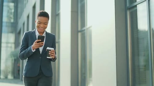 happy smiling business black woman walking on city street with mobile phone and cup coffee. African