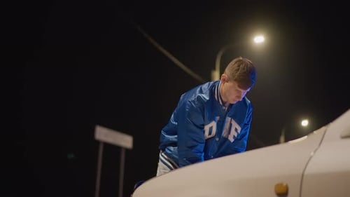 Night Roadside White Driver Inspects Hood Under Streetlight Blue Varsity Jacket Tense Expression