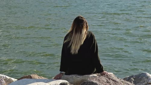 Blond Haired Woman Sitting On Stones On The Beach Looking At The Sea
