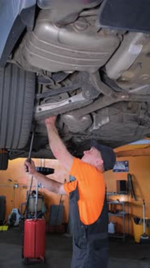 Vertical Video of a Young Mechanic in a Uniform Working in a Car Repair Shop with a Lifted Car