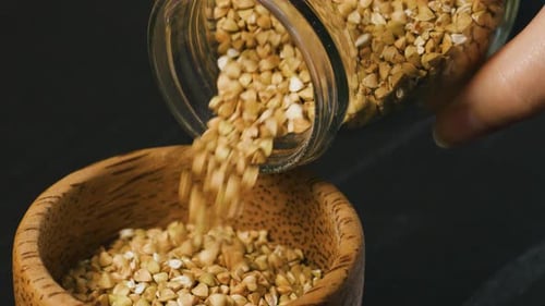 Pouring Raw Whole Grain Green Buckwheat Seeds From Glass Jar Into a Wooden Cup
