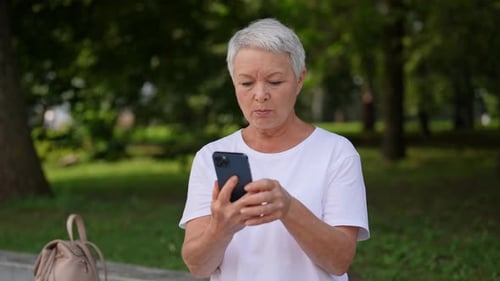 Happy European Grayhaired Older Female Sitting on Bench in City Park and Using Mobile Phone Taking