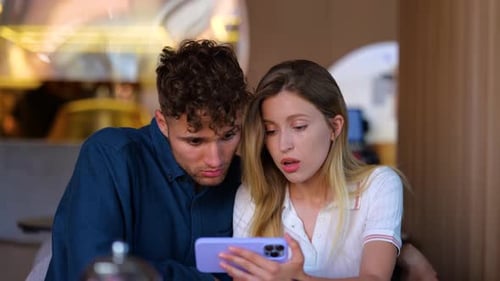 Close Up of Young Couple Man and Woman Sitting in Restaurant at Table and Looking at Smartphone