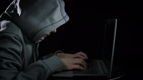 Hooded Person Typing on Laptop in Dark Room