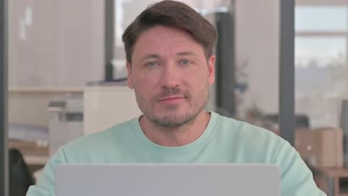 Nahaufnahme eines Mannes, der bei der Arbeit am Laptop in die Kamera schaut