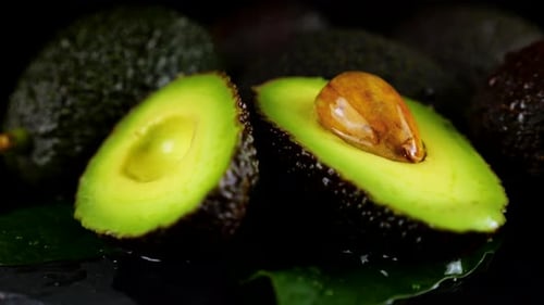 Drops of water fall on rotating avocado fruits with leaves on a black background.