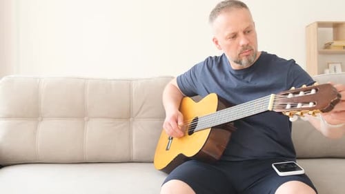 Un homme accorde une guitare acoustique assis sur un canapé