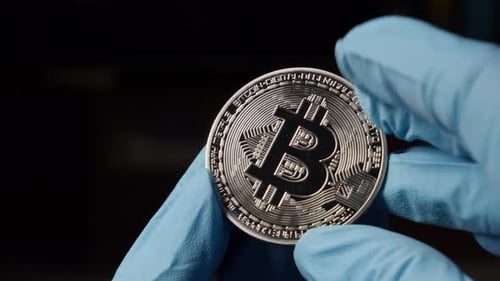 Hand With Blue Gloves Holding Bitcoin Crypto Currency Coin Against Black Background, Close Up