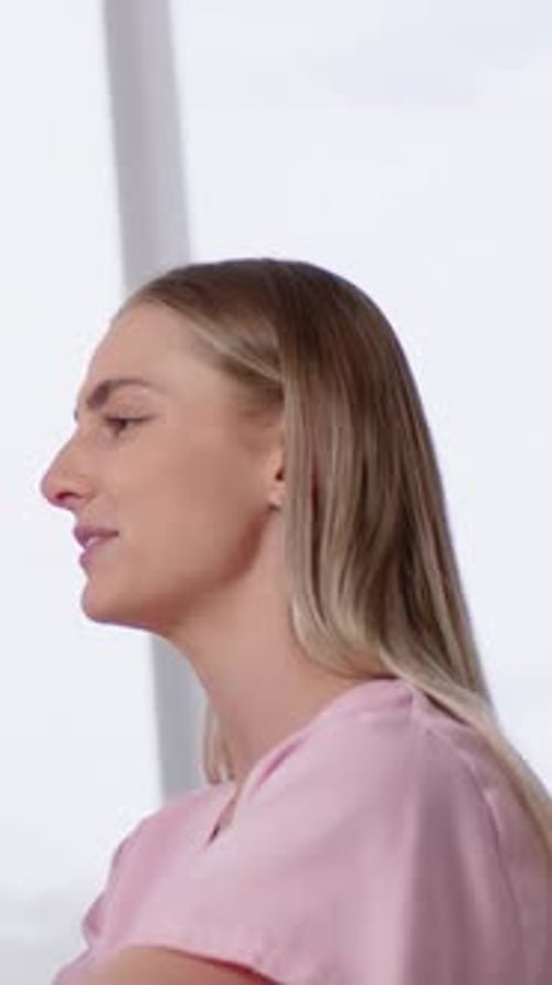 Vertical video: Speaking adult woman answering question at window, gesturing right, pink top, stud