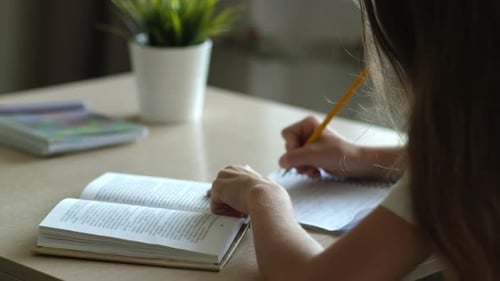 Girl Studies with Textbook, Writes on Paper at Table