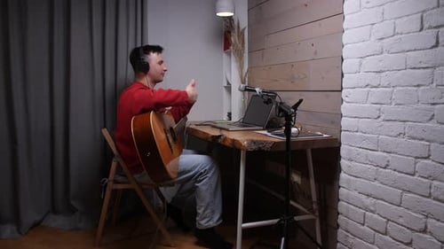 Male Musician Wearing Headphones Giving an Online Guitar Lesson From a Home Studio Smiling and