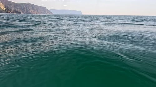 Sea Water Surface Camera Flies Over the Calm Azure Sea with Volcanic Rocky Shores on Background