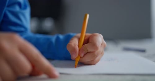 Closeup of a Child in a Blue Sleeve Drawing with a Yellow Pencil on White Paper at a Table Showing
