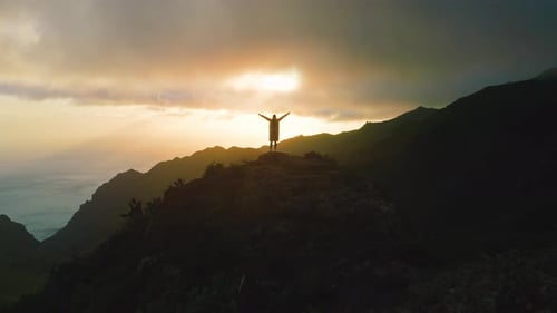 Woman Stands at Top of Mountain Over Ocean Coastline with Raised Hands in Cold Winter Weather