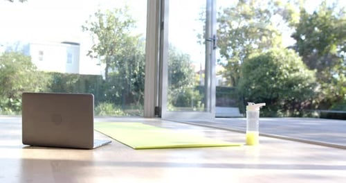 Close up of yoga mat, laptop and bottle with juice on floor at sunny home, slow motion
