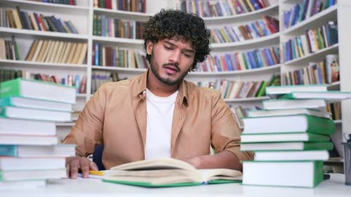 Tired student studying by reading books, taking notes in campus library space.
