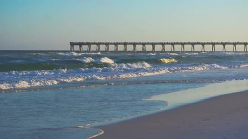 Slow Motion waves roll in at the beach with a fishing pier in the background at sunset