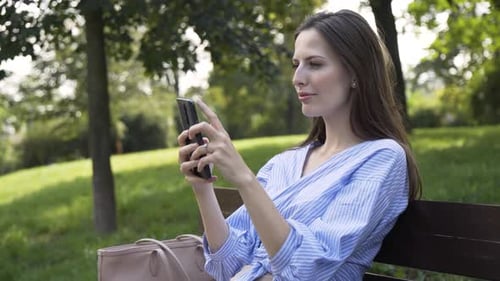 A Young Caucasian Woman Works on a Smartphone with a Smile As She Sits on a Bench in a Park