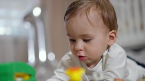 Lovely baby plays with toys lying on belly. Adorable toddler totally absorbed by the game. Close up.