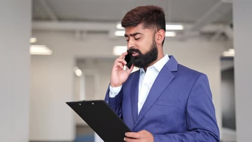 Focused Businessman Talking on Phone, Holding Clipboard in Office