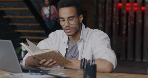 Intelligent Young Businessman Reading Book Sitting at Desk in Loft Style Coworking Center