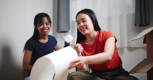 Two Asian siblings sit on the floor and help assemble a floor lamp in their new home.