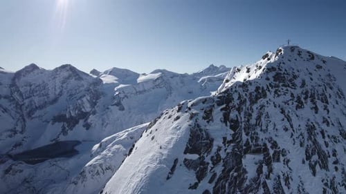Aerial panning shot of snow-covered rocky mountains with summit cross on peak in sunlight