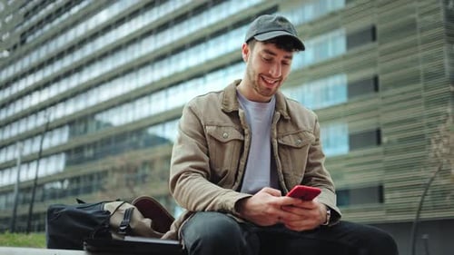Young Man Using Phone, Sitting near Urban Building