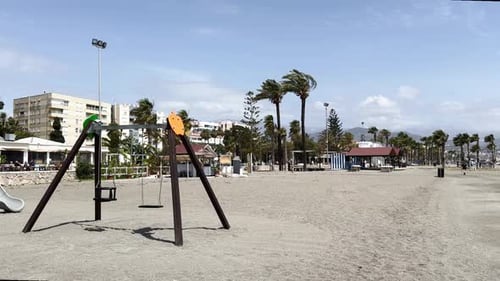 Ein verlassener Spielplatz am Strand mit Schaukeln und Rutschen, umrahmt von einer Küstenstadt im Hintergrund