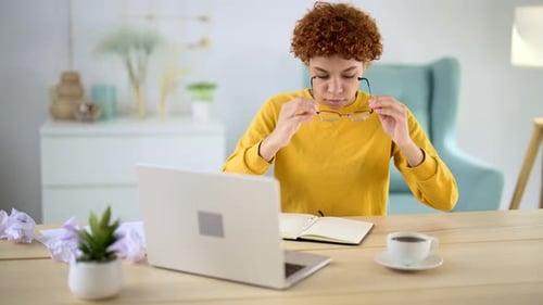 Young Woman Sits at Workplace in Home Office in Front of Laptop Screen and Looks Down at Desktop