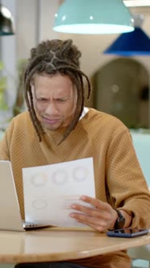 Man lifting printed charts and comparing to laptop screen for business analysis in coworking lounge