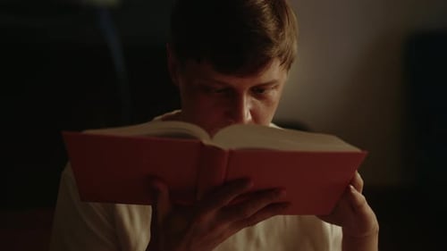 Closeup of Relaxed Young Man Enjoying Reading Interesting Book at Home in Dark Room Warm Light