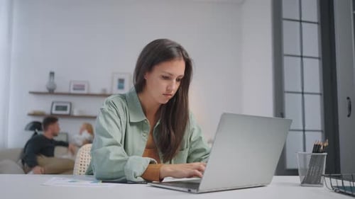 A Business Woman Works Remotely at Home on a Laptop and Shows Graphs on a Webcam While the Family is