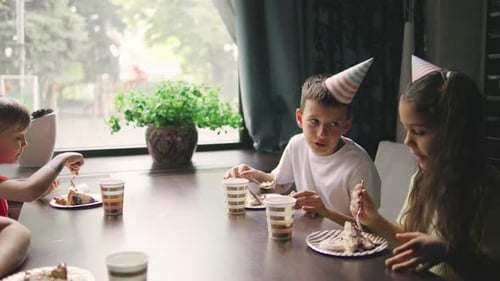 Children Have Fun Eating Birthday Cake in Festive Caps Talking and Laughing in a Cozy Festive