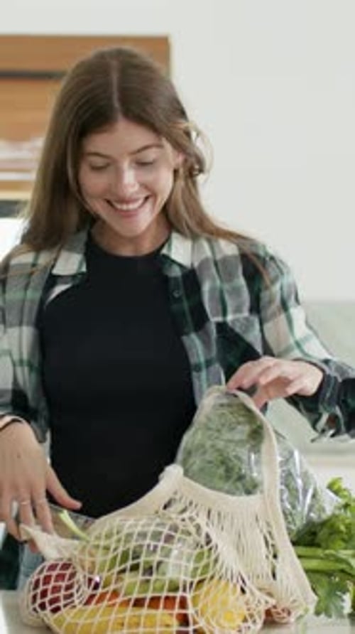 Vídeo vertical: Mujer que regresa vestida a cuadros, desempaca una bolsa de malla y comprueba las verduras envueltas en el mostrador