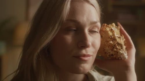 Femme blonde tenant une coquille à l'oreille et imaginant le son de l'océan à la maison