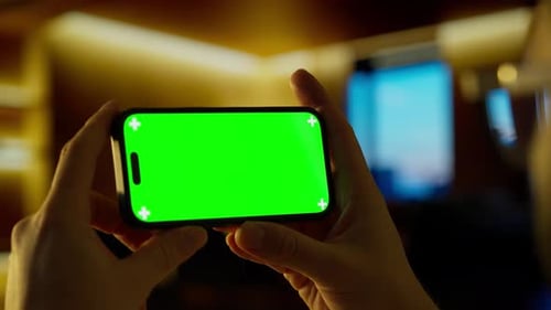 Hands Holding Modern Smartphone with Vivid Green Screen Mockup in Dimly Lit Interior Showcasing