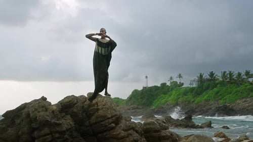 Godlike Gender Fluid Person of Color in Luxury Dress Jewelry Stands on Cliff Peak at Scenic Ocean